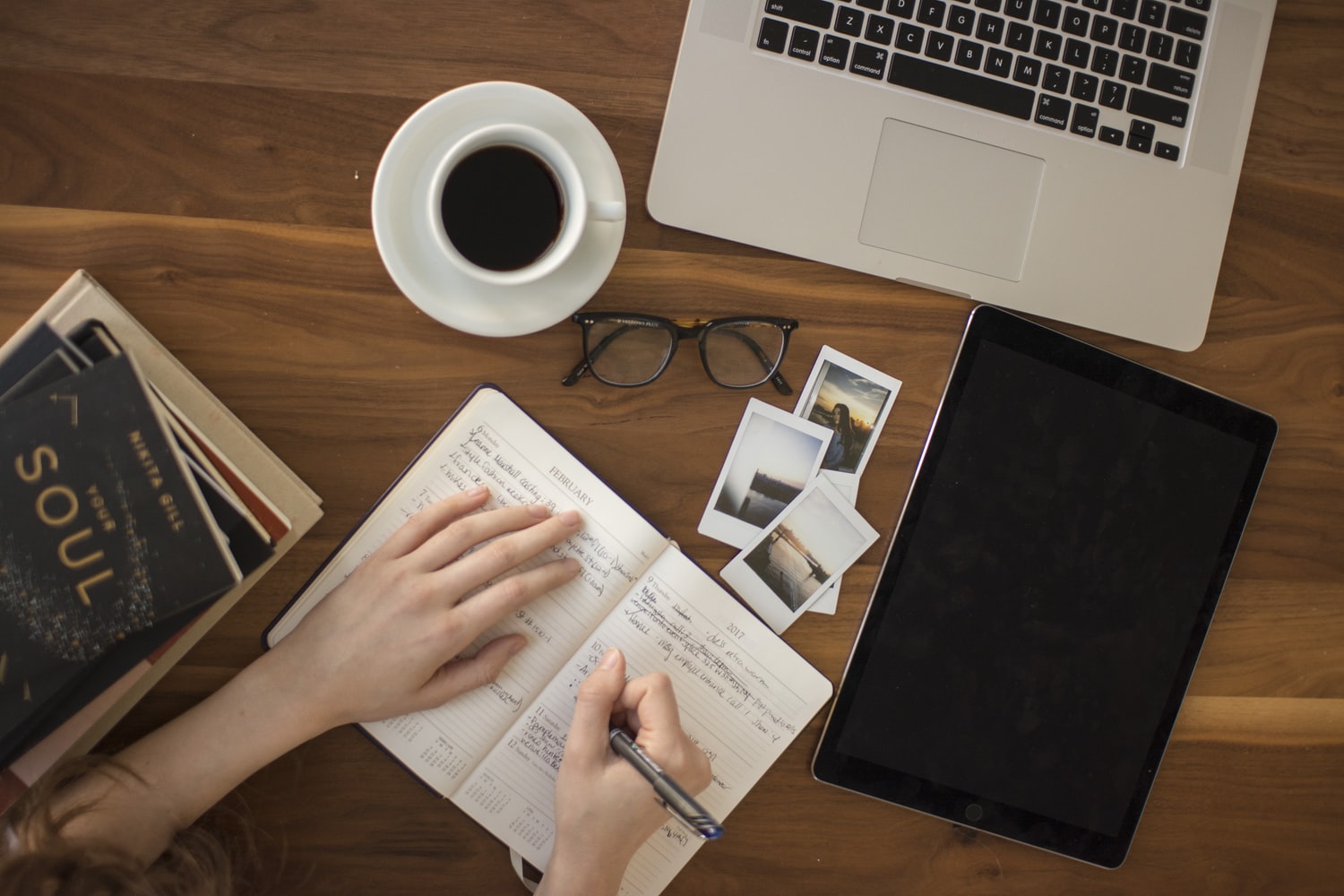 flat lay image of woman writing in notebook with coffee cup, glasses and open laptop surrounding her hands