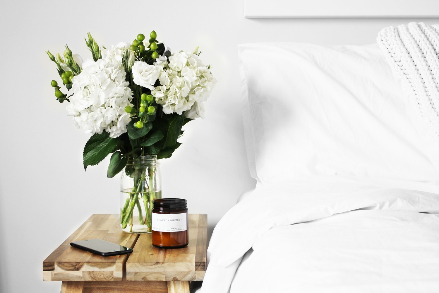 image of bed with white bedding with wooden nightstand with white flowers charging iphone and a brown glass candleholder