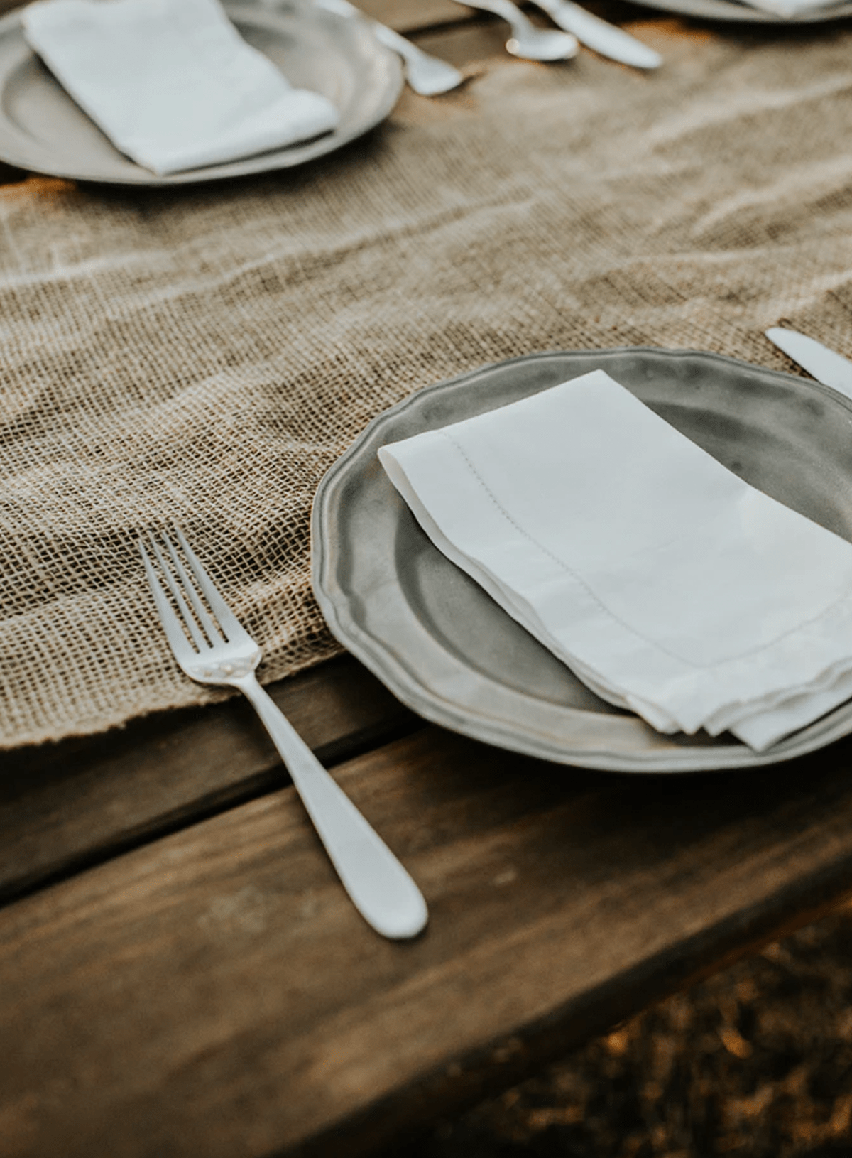 a rustic wooden table set with a burlap runner, pewter flatware, a grey scalloped edge plate and a white linen hemstitched napkin