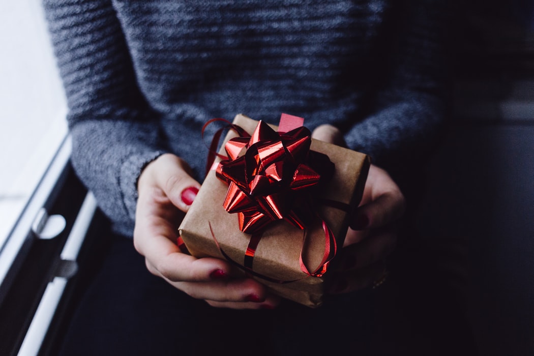 image for holiday gift guide 2019 of person with red fingernail polish holding a small package wrapped in brown paper with a red shiny ribbon on top