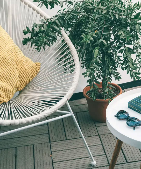 image of a white mid century acupulco chair on a grey deck with a yellow pillow, a potted tree sits next to it