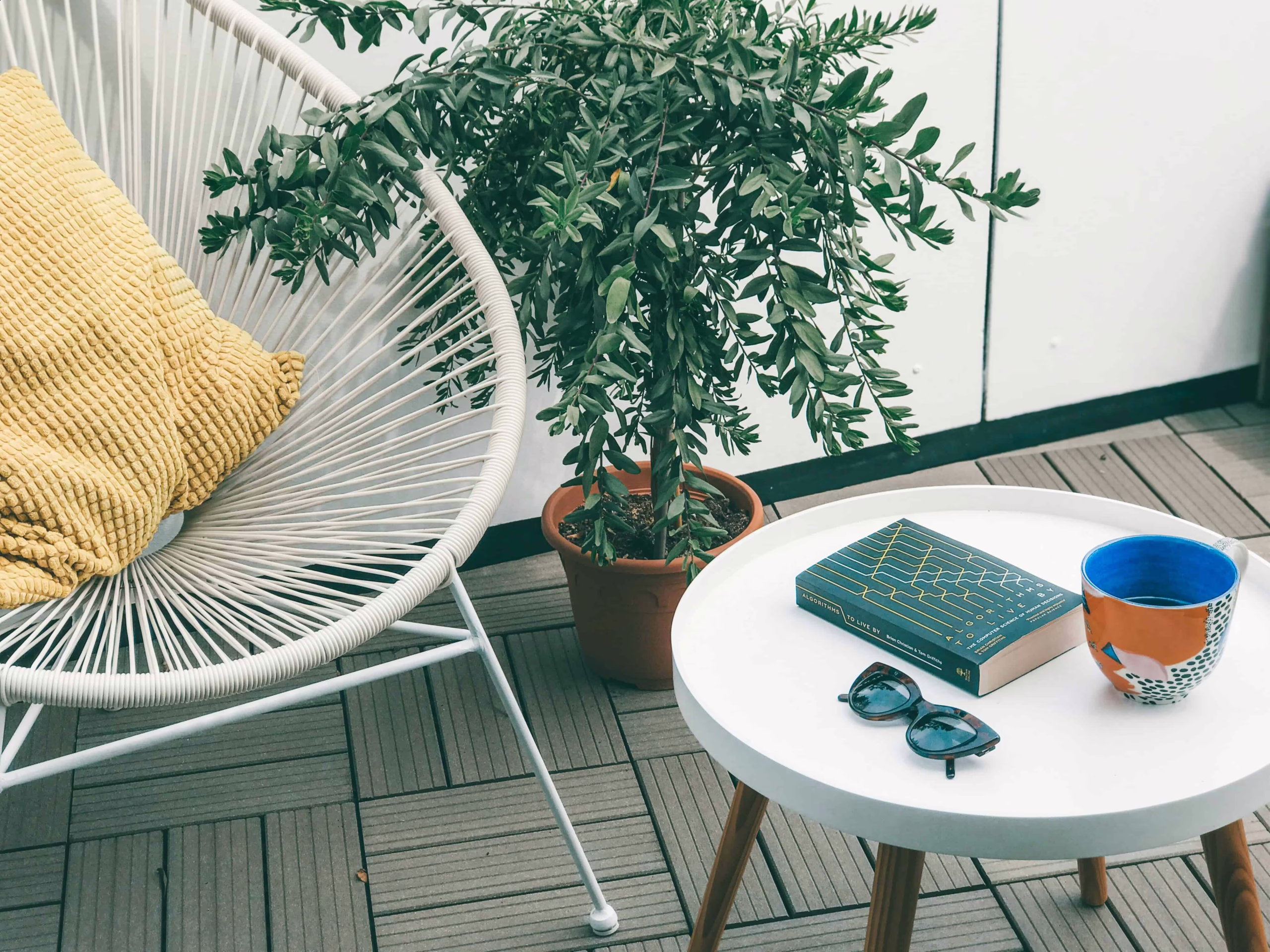 modern white patio chair with a yellow pillow next to a white side table with wood legs on a balcony with grey faux wood tile floor