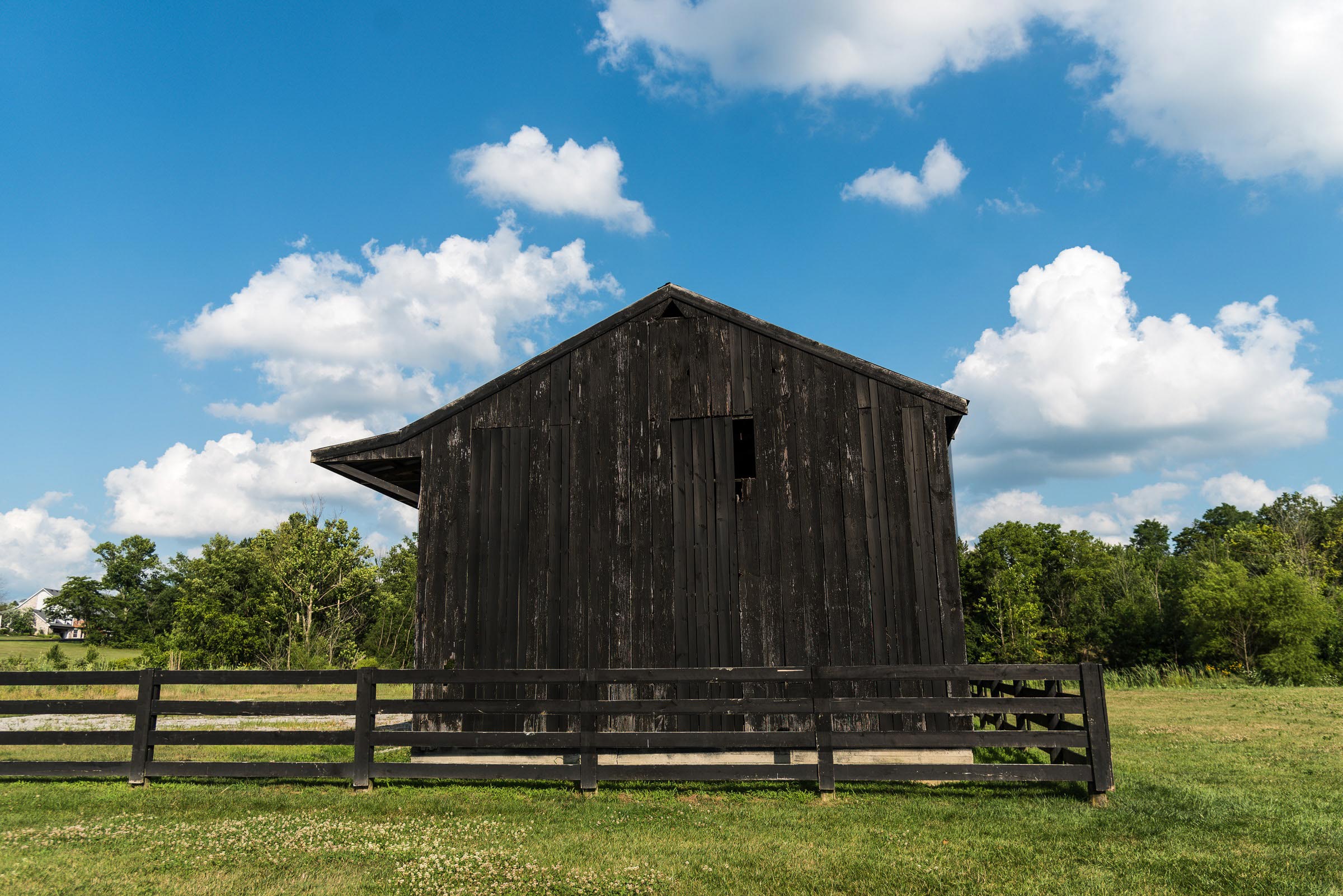 photo of a dark farm building with dark siding and a dark wood fence surrounded by green grass and blue sky