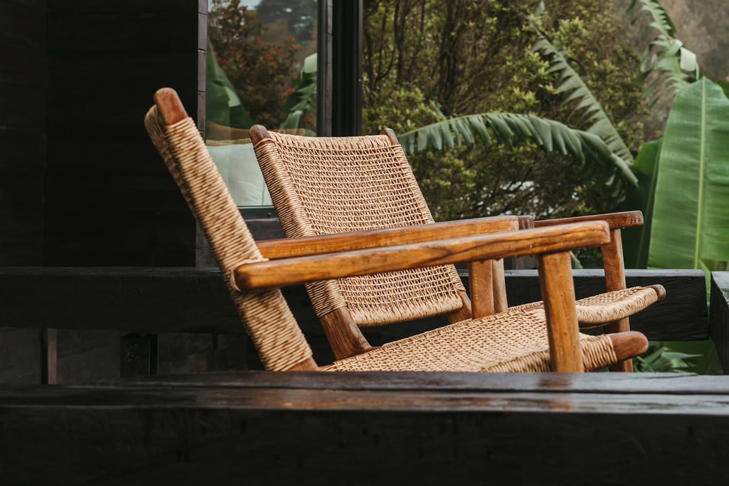woven and wood chairs on a patio in front of a house with black siding surrounded by green forest and tropical plants